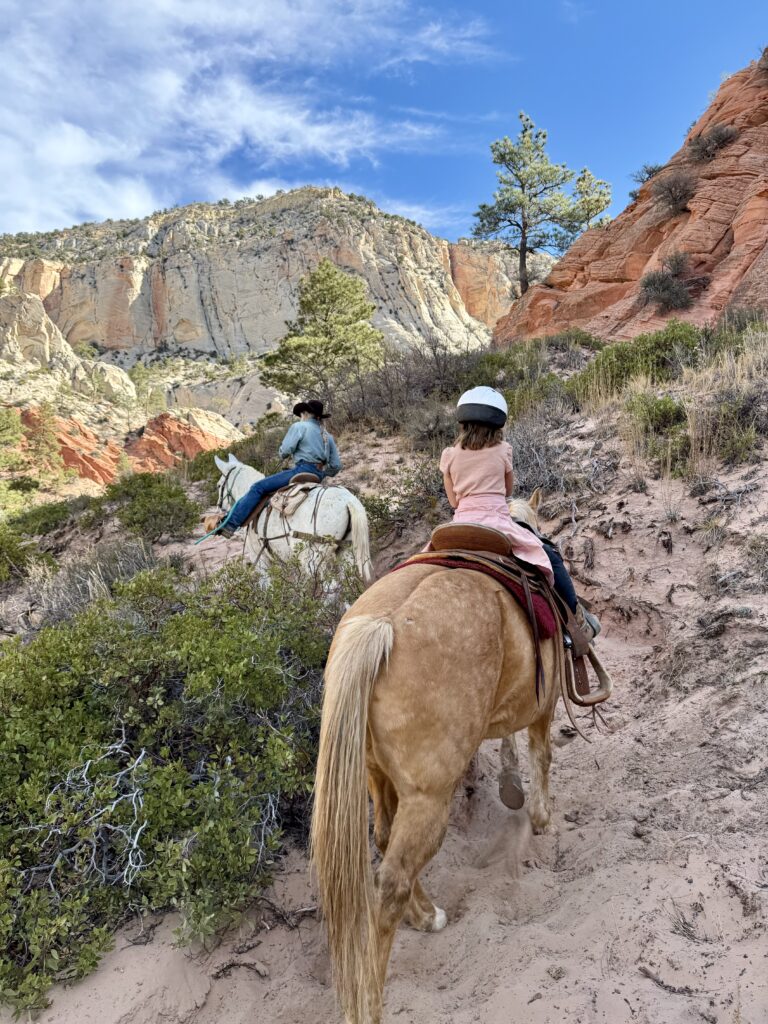 Two riders on horses navigate a rocky trail surrounded by colorful cliffs and greenery.